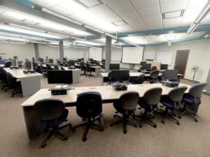 image showing classroom space with 2 retractable projection screens, 5 white boards, instructor podium, and seating with computers for 25 students