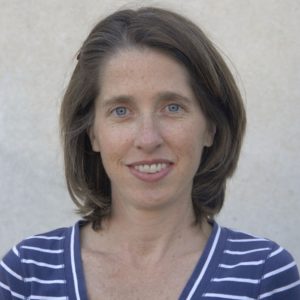 Young woman with medium brown hair smiling at camera. She is wearing a blue and white stripped shirt