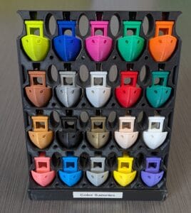 Small, 3D printed boats in a rack demonstrating the colors available to use. These colors are, from top to bottom and left to right: bright green, blue, bambu green, orange, brown, silver, light gray, red, mistletoe green, gold, black, bronze, beige, jade white, pink, cyan, gray, yellow, and purple.