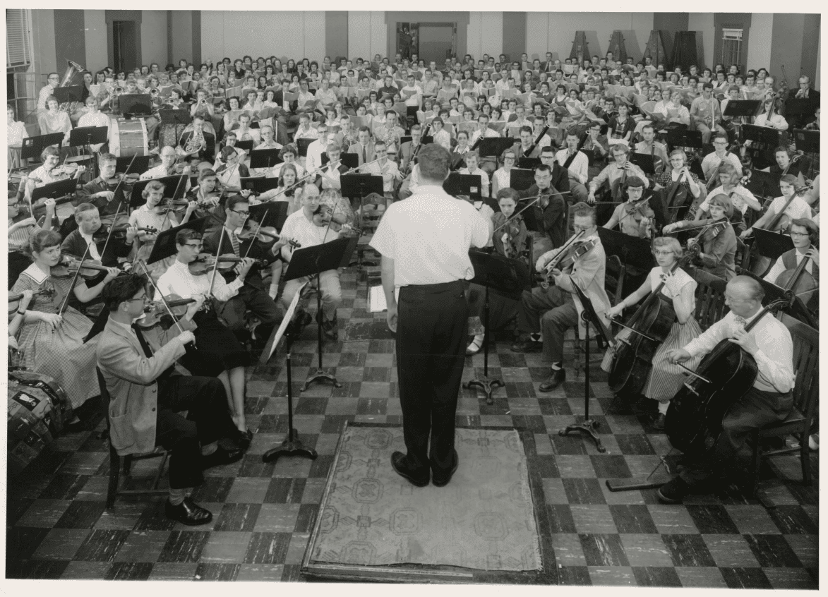 Dixon conducting a very large symphony orchestra in a practice hall. His back is to the camera and he faces the orchestra.