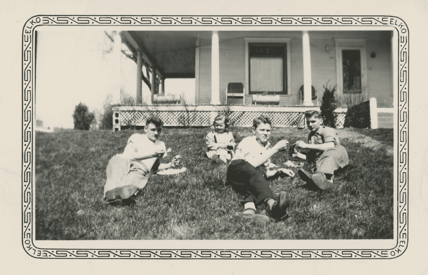 Black and white photo of four young children relaxing on the front lawn of a modest white home in Guthrie Center, Iowa.