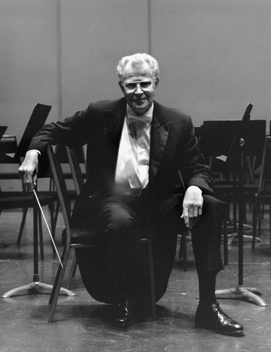 Black and white photo of older Dixon in the 1990s seated in the first violins section of the orchestra. Chairs are empty except for him.