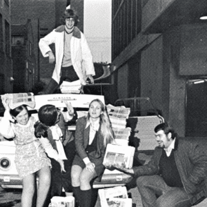 Five students sit near the trunk of a vehicle in an alleyway. They are surrounded by bundles of student published newspapers and are smiling. The photo is in black and white.