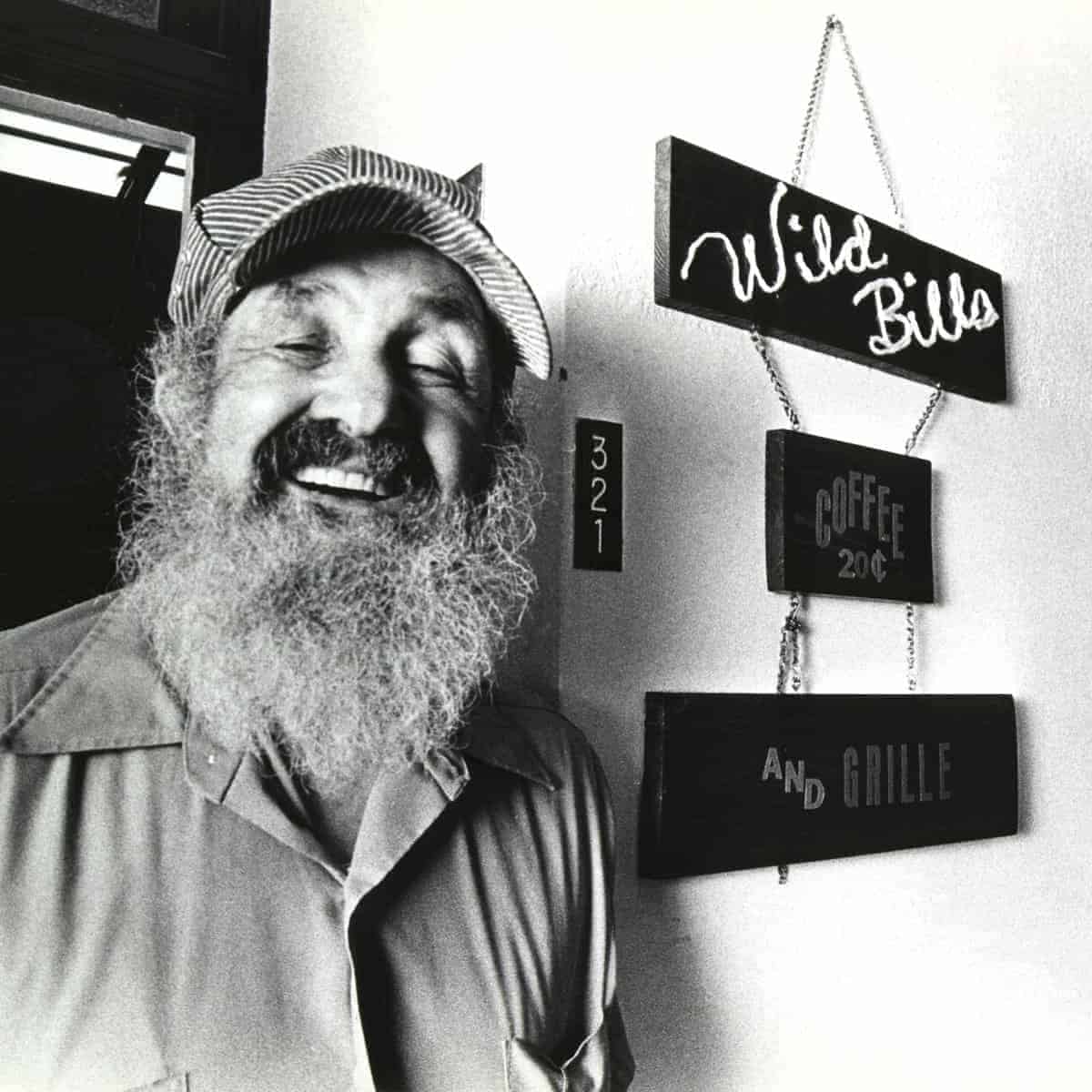 Black and white photo of Bill Sackter, an older man with a long gray beard, a vibrant smile, and a railroad engineer's cap. He stands next to a sign that says Wild Bill's Coffee.
